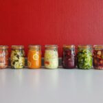 A row of vibrant, multicolored mason jars filled with assorted fermented vegetables against a red wall.