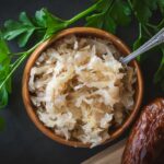 From above, a small wooden bowl is filled with tangy sauerkraut, its textured appearance offering a hint of its sharp flavor. A metal teaspoon rests adjacent, ready for serving. This setting highlights sauerkraut as a quintessential garnish, perfect as a bratwurst or hotdog topping, adding a delightful crunch and zest to each bite.