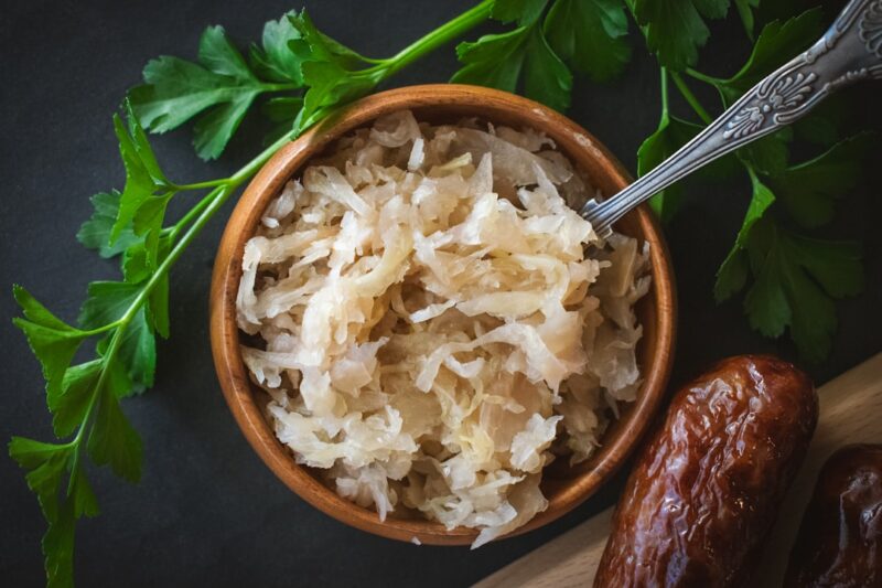 From above, a small wooden bowl is filled with tangy sauerkraut, its textured appearance offering a hint of its sharp flavor. A metal teaspoon rests adjacent, ready for serving. This setting highlights sauerkraut as a quintessential garnish, perfect as a bratwurst or hotdog topping, adding a delightful crunch and zest to each bite.