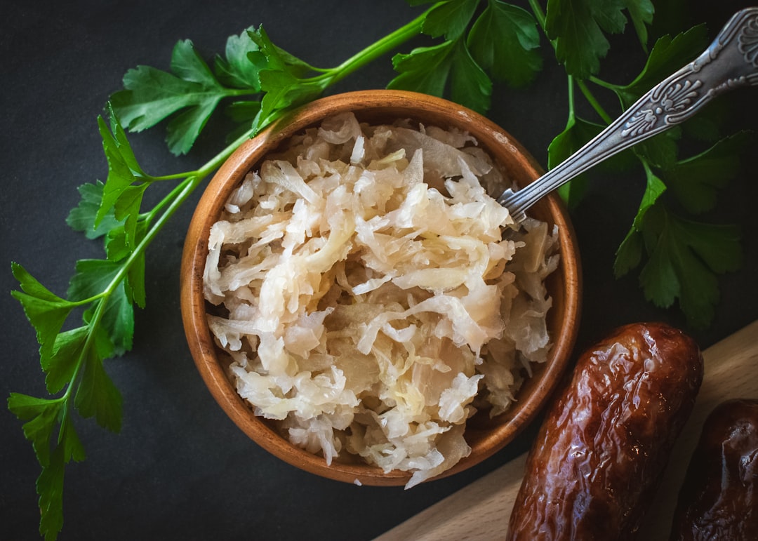 From above, a small wooden bowl is filled with tangy sauerkraut, its textured appearance offering a hint of its sharp flavor. A metal teaspoon rests adjacent, ready for serving. This setting highlights sauerkraut as a quintessential garnish, perfect as a bratwurst or hotdog topping, adding a delightful crunch and zest to each bite.