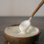From above of anonymous person stirring sour cream with spoon in wooden bowl placed on table against blurred background